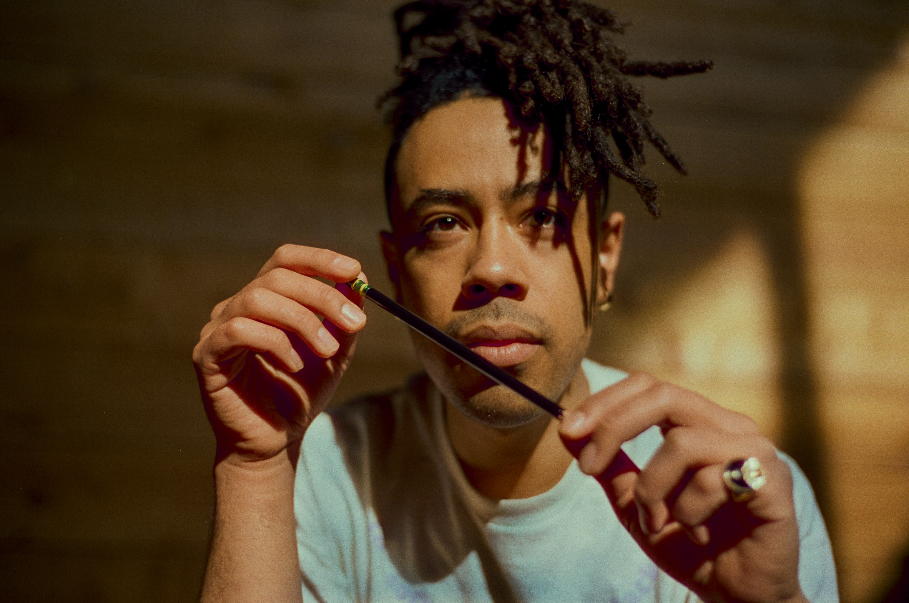 Photo of Alan Pelaez Lopez, a non-binary Black and Indigenous person with dark hair in short locs, holding a pencil with sun shining from a window